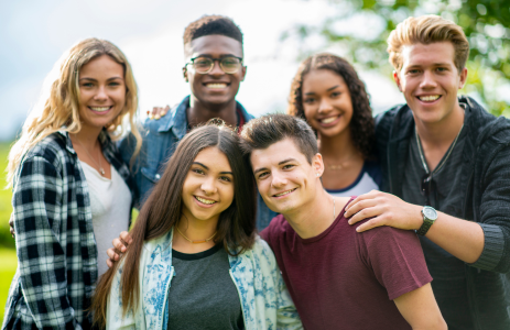 Group of teens outside, smiling into the camera. The sun is shining and there are green trees behind them.