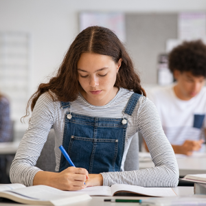 Young girl in overalls writing at a desk with her head down.