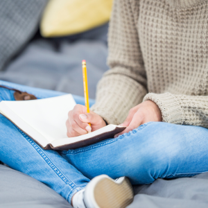 Teen's torso and legs sitting crossed on a bed, writing in a notebook