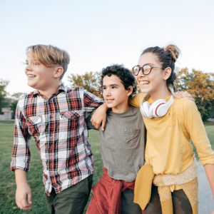 Three kids walk with their arms around each other in the late afternoon, smiling and talking looking away from the camera